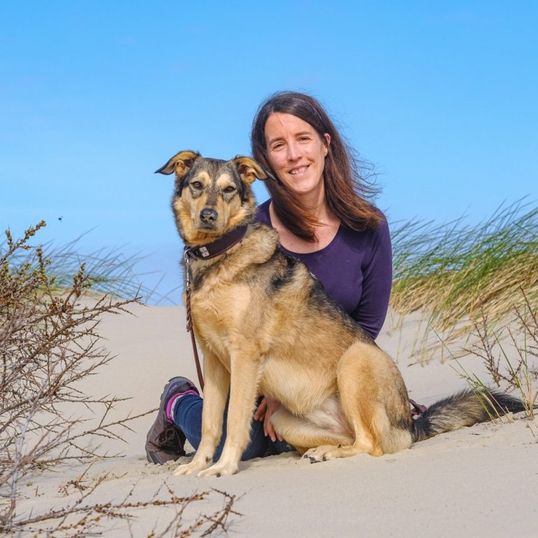 Eine Frau sitzt im Sand neben einem Hund bei sonnigem Wetter.