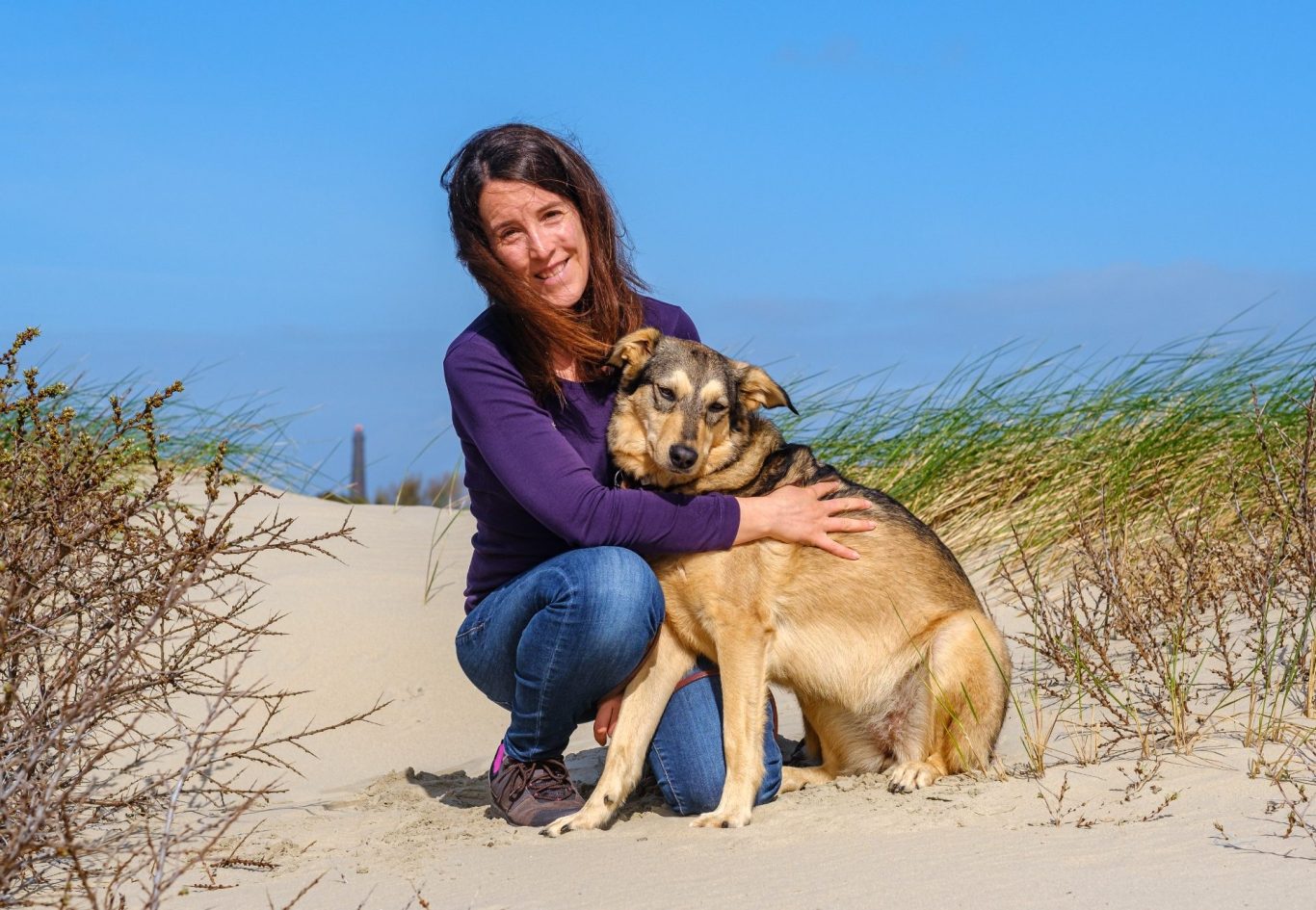 Frau kniet im Sand und umarmt ihren Hund, umgeben von Gras und blauem Himmel.